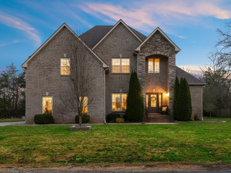 Two-story brick and stone home with a lit arched entry and evergreen shrubs at dusk at 4424 Thoroughbred Lane, Murfreesboro, Tennessee